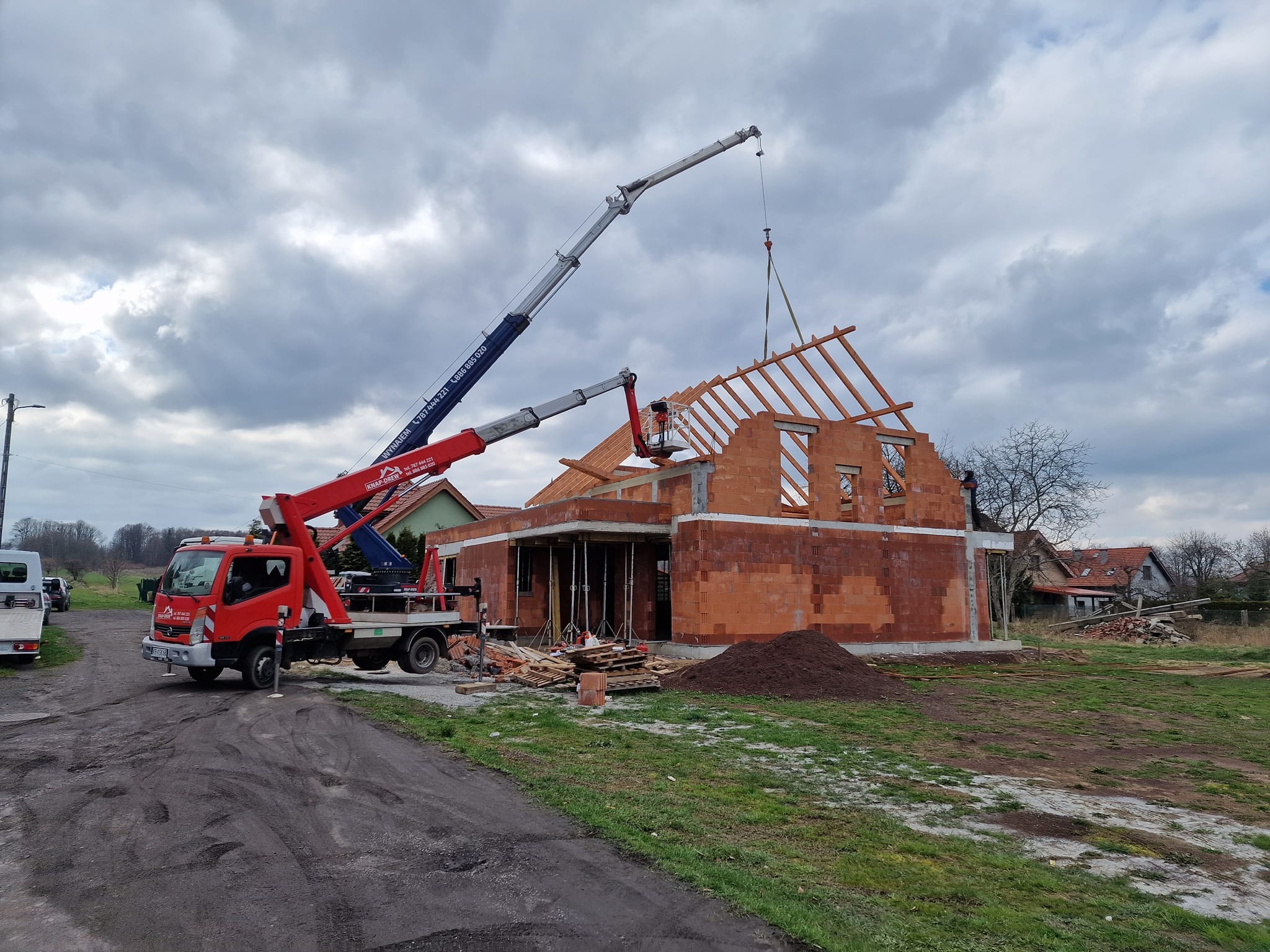 Budowa domu jednorodzinnego: czerwona cegła, drewniana więźba dachowa w trakcie montażu. Dwa dźwigi z platformami roboczymi, pochmurne niebo w tle.