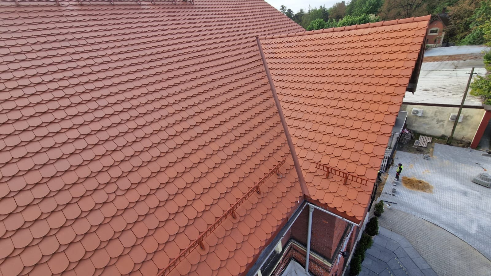 Czerwony dach pokryty dachówką karpiówką, widoczny z góry, z elementami systemu przeciwśniegowego. W tle drzewa i budynki.