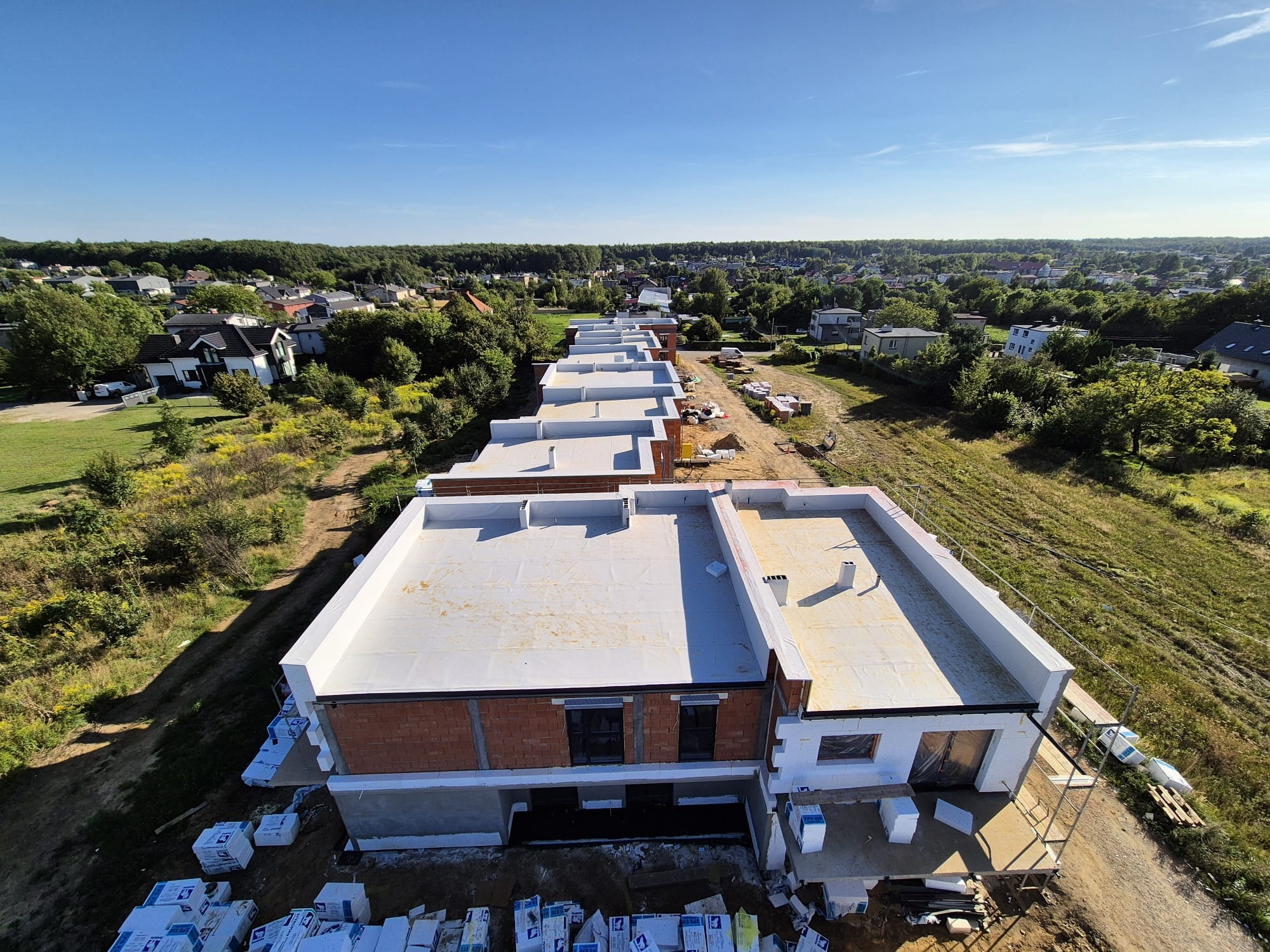 Widok z góry na szereg budynków z płaskimi dachami w trakcie budowy. Ceglane ściany, białe wykończenia, materiały budowlane na ziemi. W tle zabudowa jednorodzinna i zieleń.