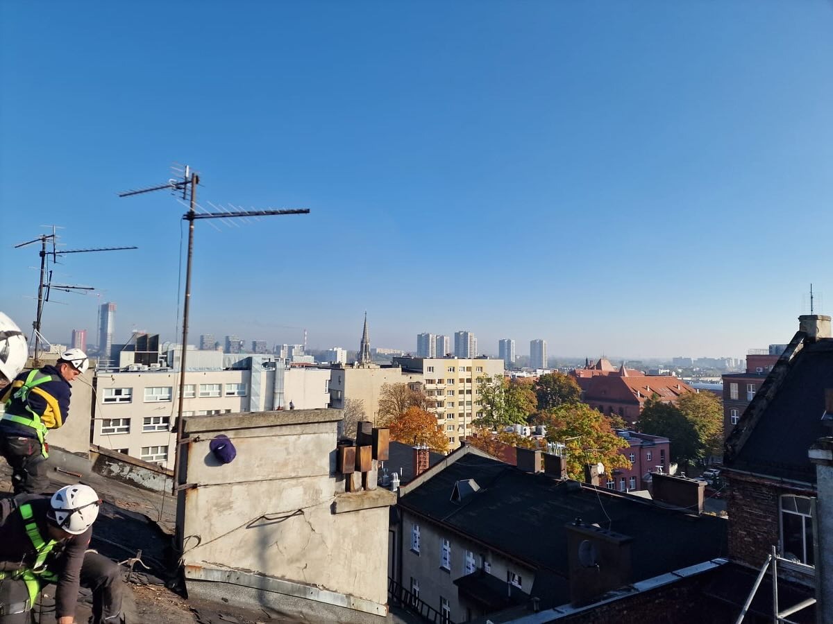 Praca na dachu kamienicy w tle miejskiej panoramy. Dwóch dekarzy w kaskach i uprzężach, anteny na dachu, błękitne niebo. Praca na wysokościach w słoneczny dzień.