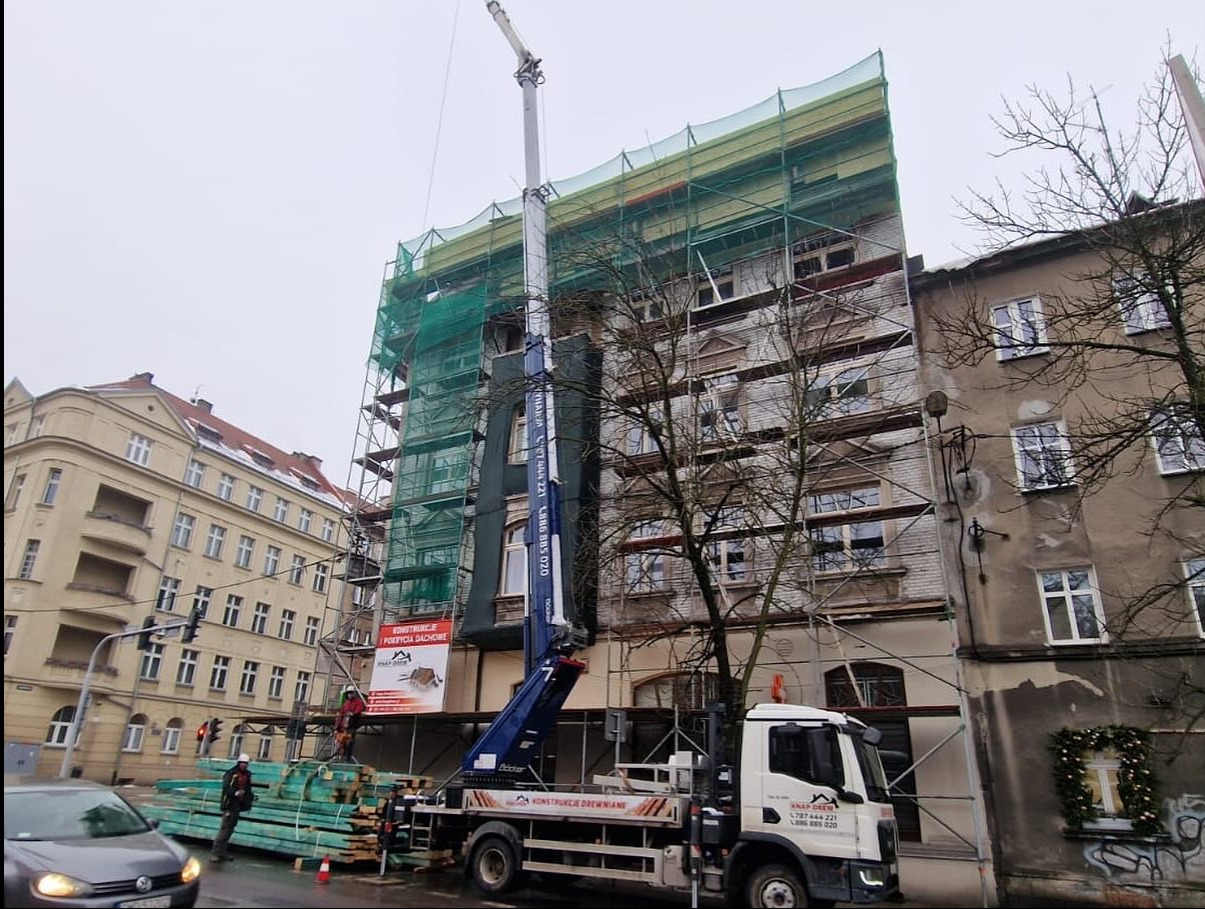 Budynek w rusztowaniach, zabezpieczony siatką. Na pierwszym planie dźwig z materiałami budowlanymi. Widoczni pracownicy w kaskach. Prace dekarskie w toku.