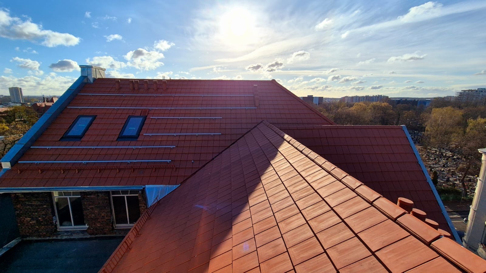 Czerwony dach z dachówką ceramiczną, widoczne okna dachowe i elementy montażowe paneli solarnych na tle błękitnego nieba z chmurami i słońcem.