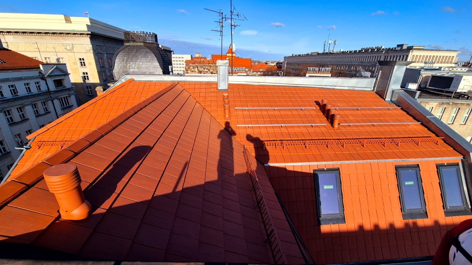 Czerwony dach z dachówki ceramicznej w miejskiej zabudowie, widoczne okna dachowe, komin i cień osoby. Ujęcie z góry, w tle budynki.