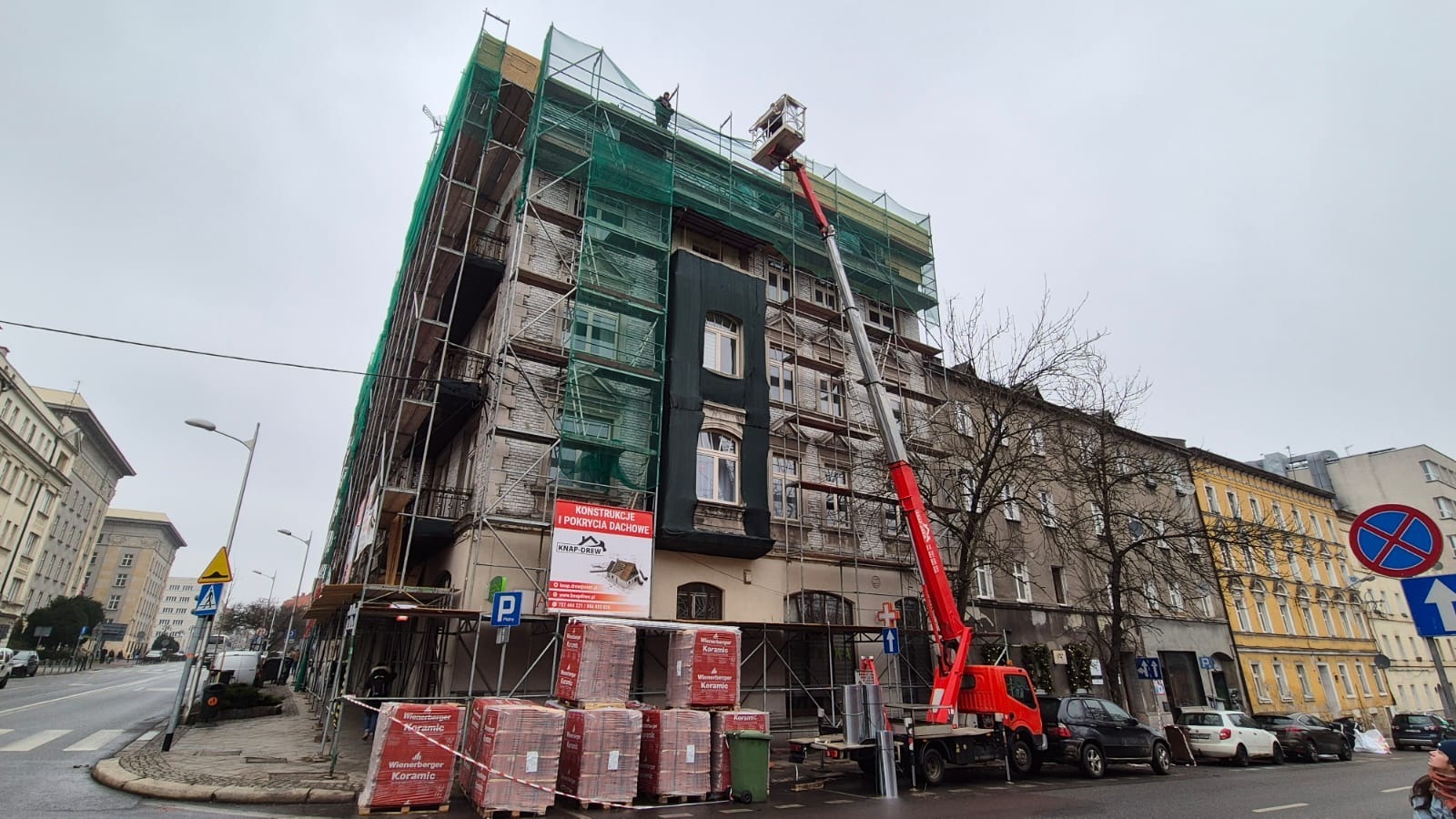 Budynek w rusztowaniach z zieloną siatką, widoczny dźwig i palety z czerwoną dachówką ceramiczną. Na budynku baner informujący o konstrukcji i pokryciu dachowym.