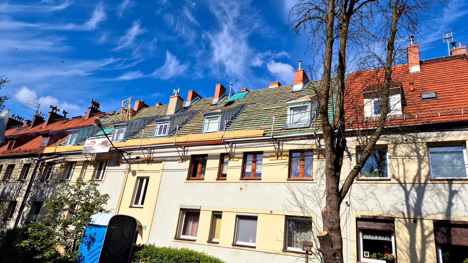 Wymiana dachówki na budynku wielorodzinnym. Widoczne rusztowania i częściowo nowy dach. Błękitne niebo z chmurami w tle. Przenośna toaleta na pierwszym planie.