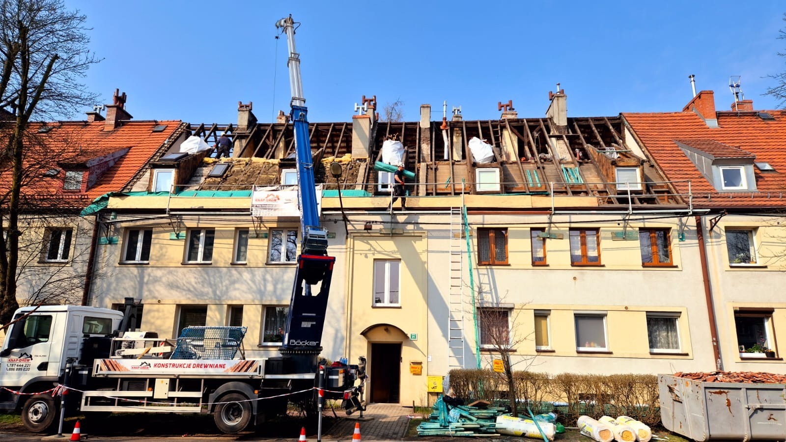 Demontaż starego dachu na budynku wielorodzinnym. Prace dekarskie w toku, widoczny dźwig i pracownicy na dachu. Częściowo odkryta więźba dachowa.