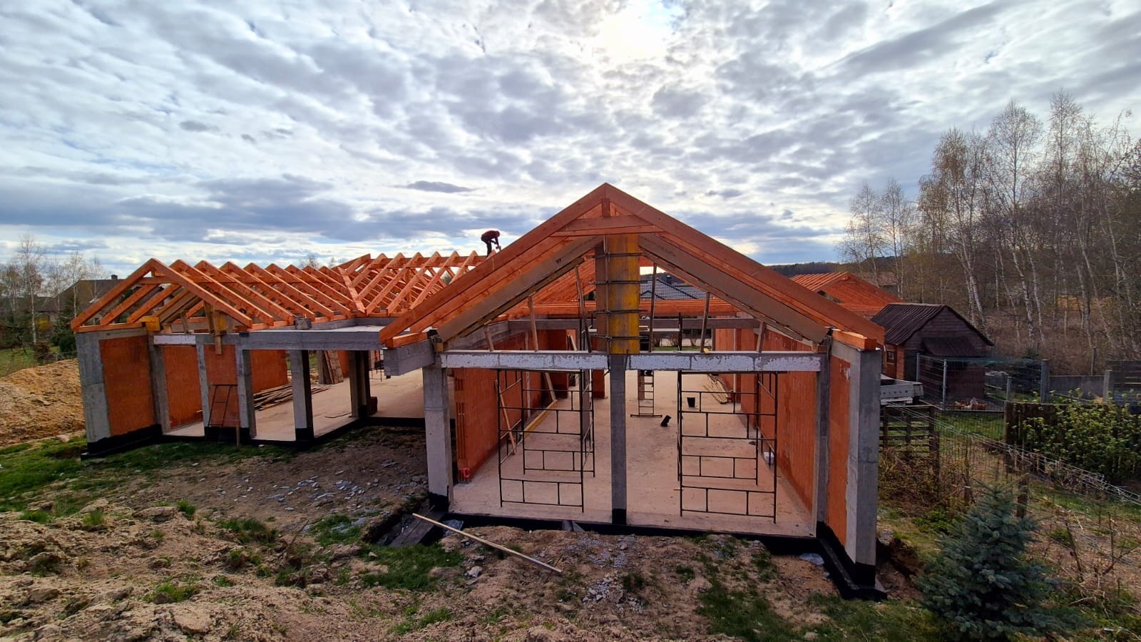 Wznoszenie konstrukcji domu z drewnianą więźbą dachową. Widoczny pracownik na dachu, ceglane ściany i betonowe słupy. Tło stanowią drzewa i pochmurne niebo.