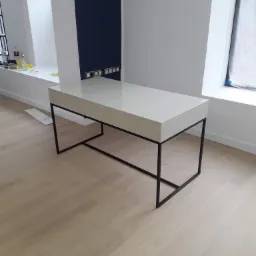 Białe, lakierowane biurko na metalowym stelażu o minimalistycznym designie, ustawione na jasnej drewnianej podłodze. W tle białe ściany z granatowym akcentem i gniazdkami elektrycznymi.