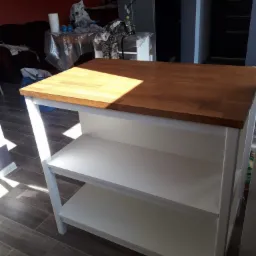 Kuchenny stół roboczy z blatem z jasnego drewna i białymi półkami, ustawiony na podłodze z płytek imitujących drewno, z widocznym fragmentem zagraconego pomieszczenia w tle.