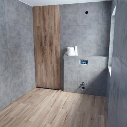 Kornel Kłosinski Usługi remontowo-wykończeniowe - Wnętrze łazienki w trakcie remontu, widoczne szare płytki ścienne, drewnopodobne płytki podłogowe, zabudowa WC ze stelażem podtynkowym, rolka papieru toaletowego i otwory na instalacje.