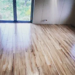 Ułożona podłoga z paneli imitujących drewno, widoczne połączenia między panelami, ściana wykończona płytą karton gips, okno z widokiem na zieleń.