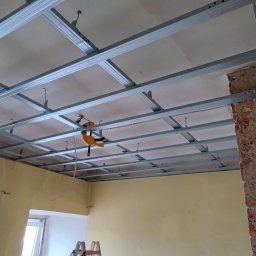 Kornel Kłosinski Usługi remontowo-wykończeniowe - Stelaż sufitu podwieszanego z profili metalowych, mocowany do surowego stropu, z widocznym starym murem z cegły po prawej stronie.