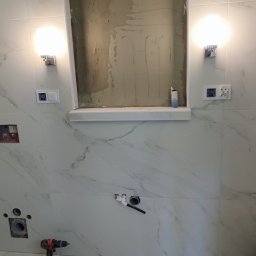 Kornel Kłosinski Usługi remontowo-wykończeniowe - Niedokończona wnęka w ścianie łazienki z marmurowymi płytkami, widoczne instalacje hydrauliczne, wiertarka akumulatorowa i lampy ścienne.