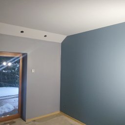 Kornel Kłosinski Usługi remontowo-wykończeniowe - Pokój po częściowym pomalowaniu, dwie ściany w odcieniach szarości, widoczne halogeny budowlane doświetlające ścianę, okno balkonowe z widokiem na zaśnieżony ogród.