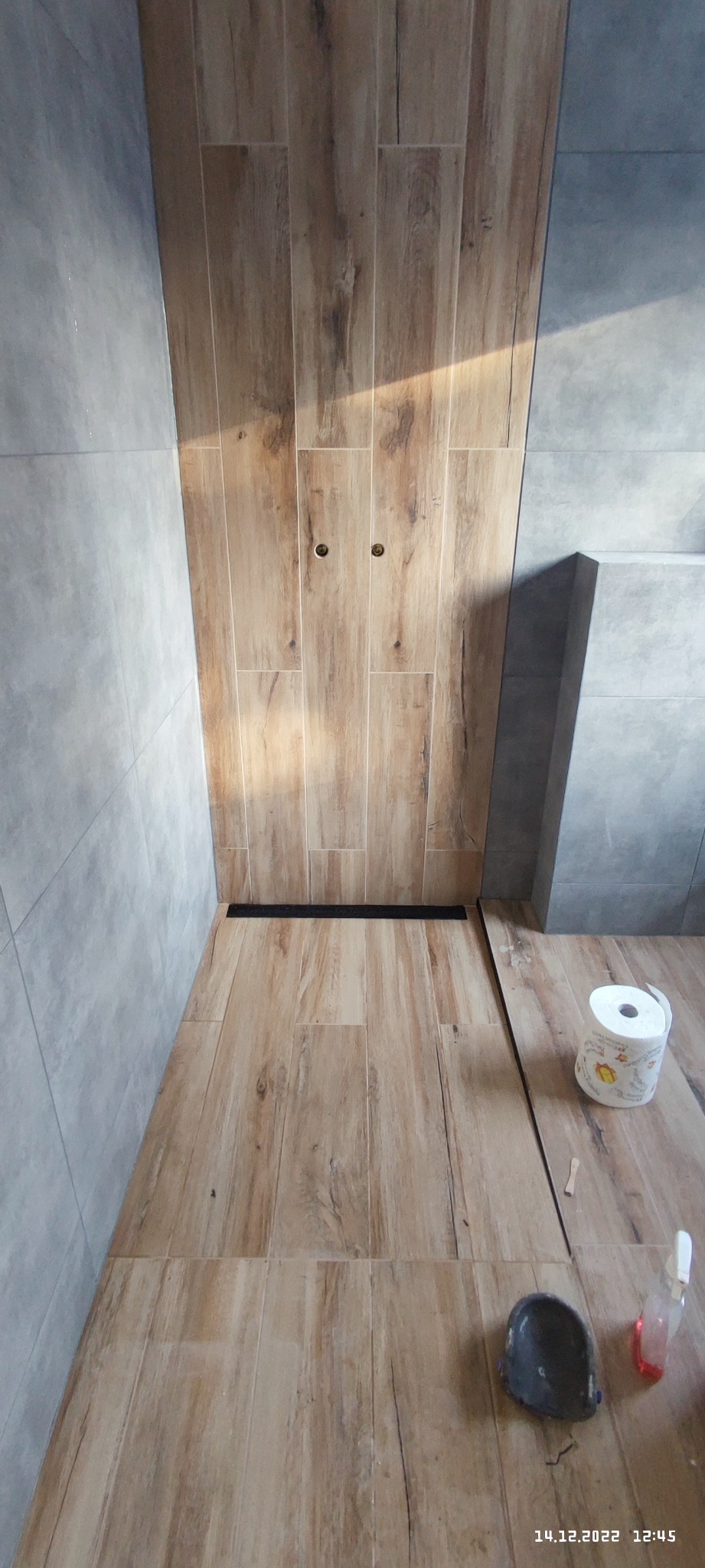 Wykończona łazienka z płytkami imitującymi drewno na podłodze i ścianie prysznica, widoczny odpływ liniowy, szare płytki na pozostałych ścianach, rolka papieru toaletowego i akcesoria do sprzątania...