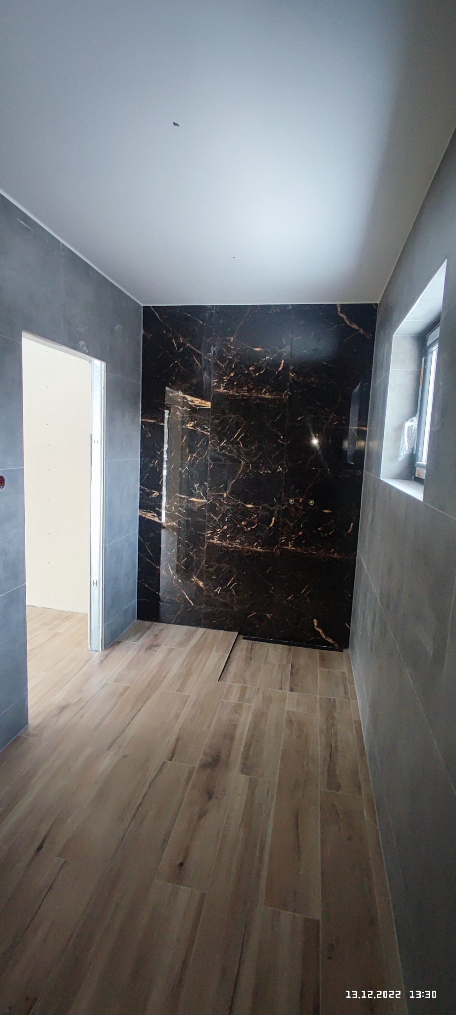 Nowoczesna łazienka w trakcie wykańczania: płytki imitujące drewno na podłodze, ściana prysznicowa z czarnych płytek z marmurkowym wzorem, szare płytki na pozostałych ścianach, widoczny odpływ...