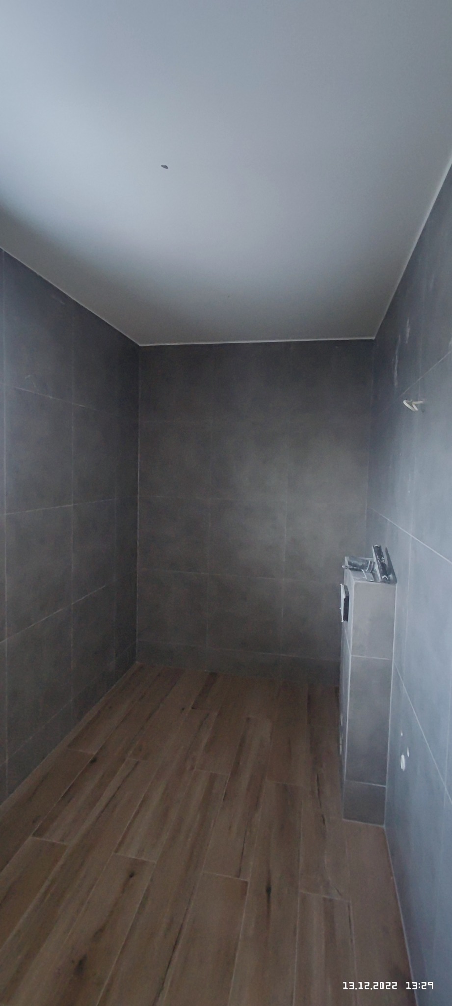 Wnętrze łazienki w trakcie remontu, z szarymi płytkami na ścianach i drewnopodobnymi panelami na podłodze, widoczny fragment zabudowy z narzędziami na górze.