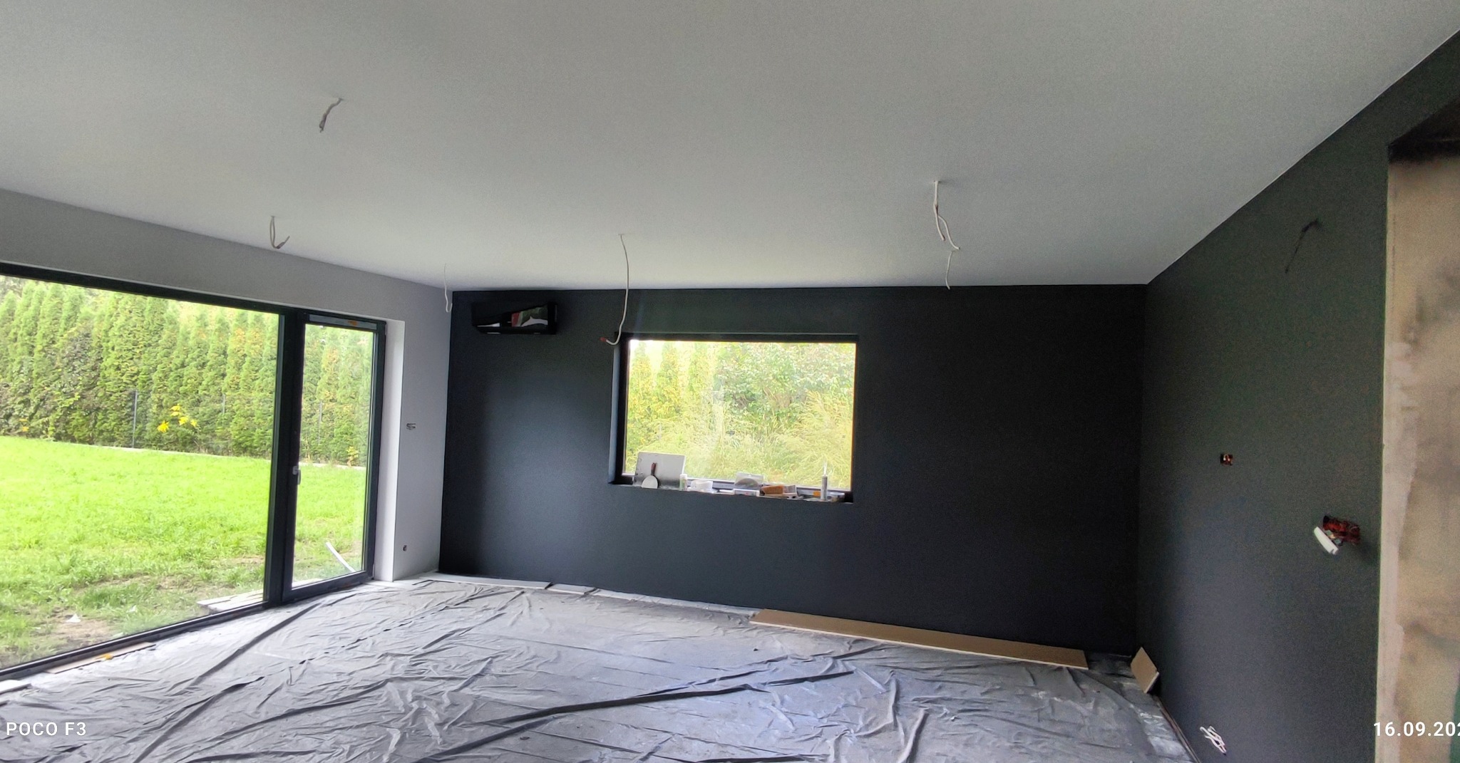 Pokój po świeżym pomalowaniu ścian na ciemny kolor, zabezpieczona podłoga folią, widoczne wystające kable i narzędzia malarskie na parapecie okna.