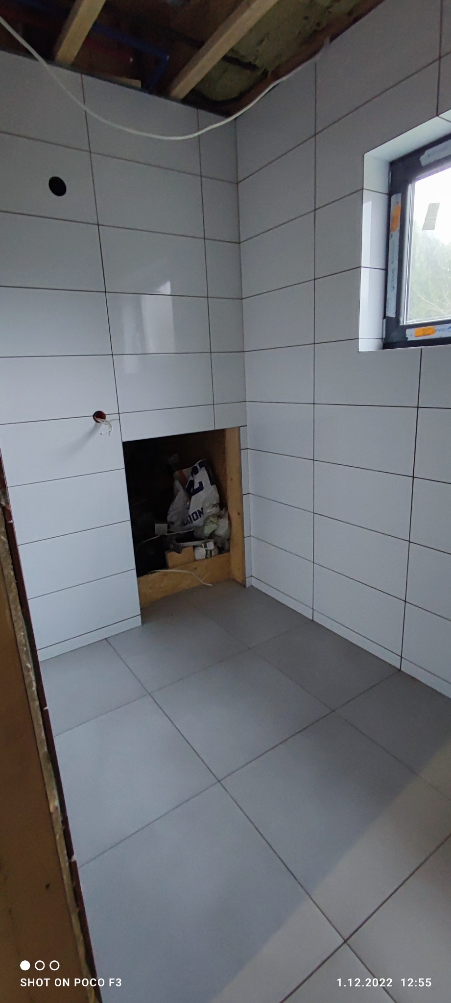 Świeżo wykafelkowana łazienka z białymi płytkami ściennymi i szarymi podłogowymi, widoczne otwory na instalacje i niezabezpieczona wnęka w ścianie z elementami instalacji.