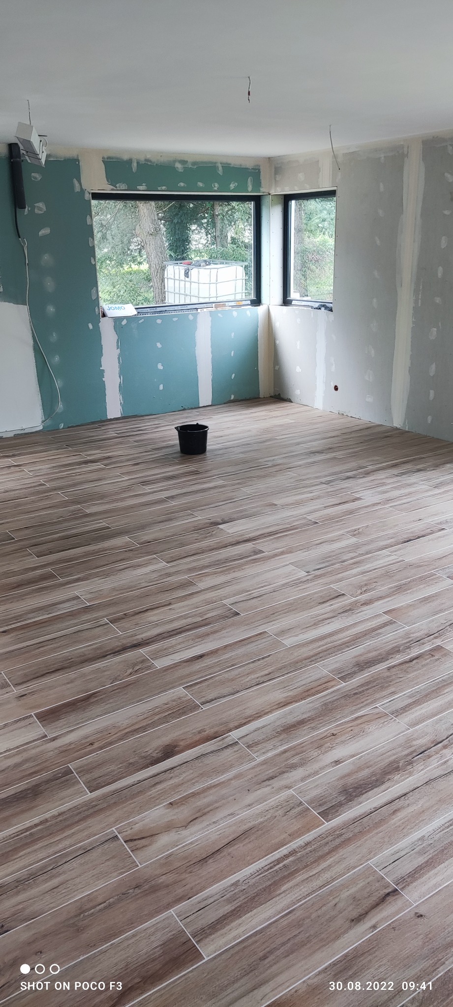 Wnętrze w trakcie remontu: położona podłoga imitująca drewno, ściany częściowo pokryte płytami kartonowo-gipsowymi, widoczne okno i czarne wiadro na środku pomieszczenia.