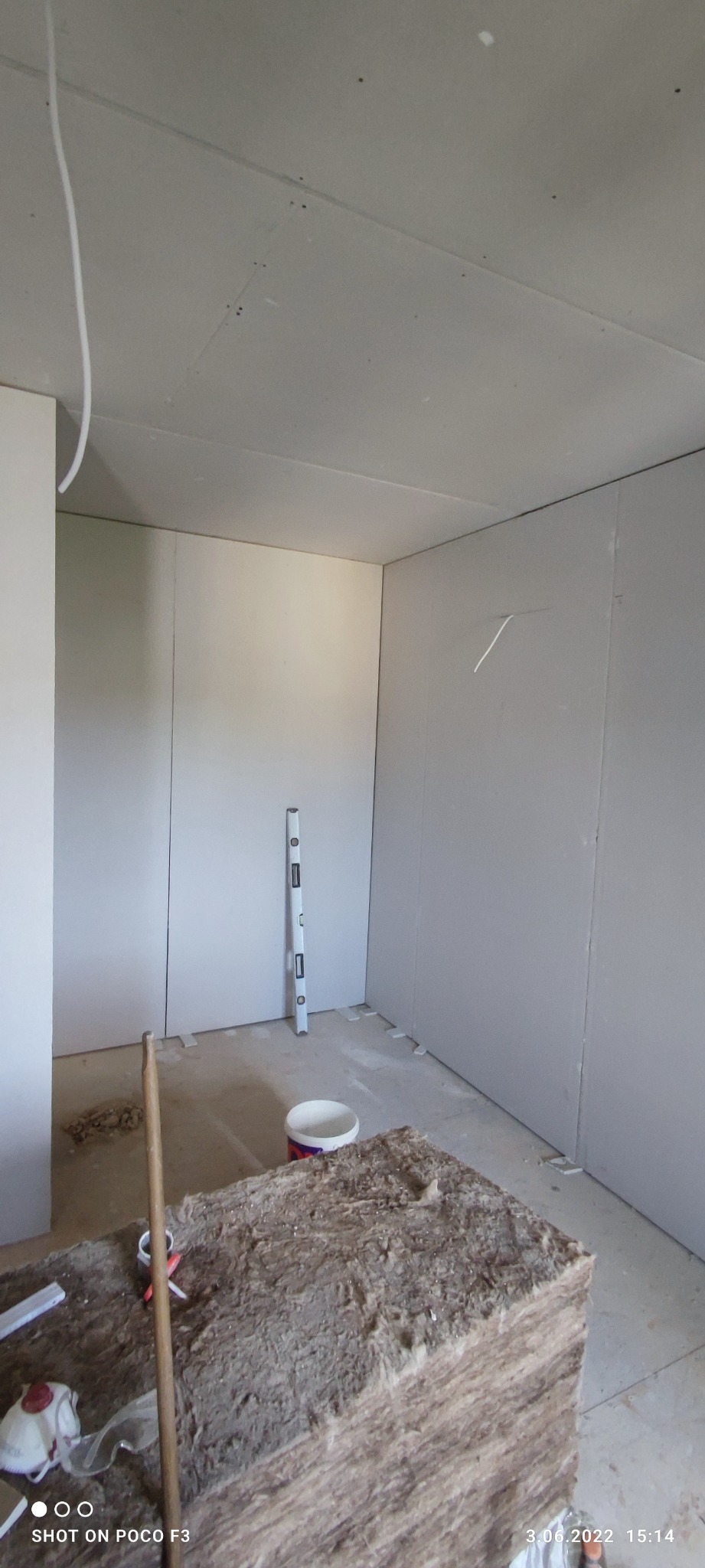 Wnętrze w trakcie prac wykończeniowych, ściany obłożone płytami kartonowo-gipsowymi, widoczny poziomica przy ścianie, paczka wełny mineralnej i wiadro farby na podłodze.