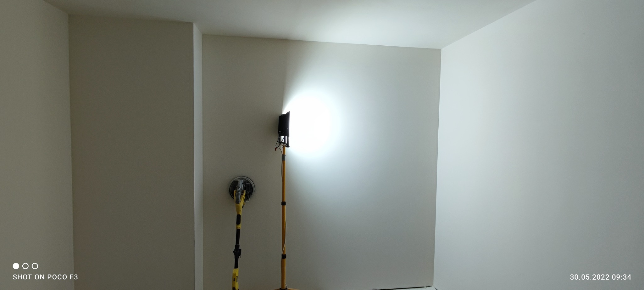 Wnętrze pokoju z wyszlifowanymi ścianami, oświetlone lampą roboczą, obok stoi szlifierka do gładzi.