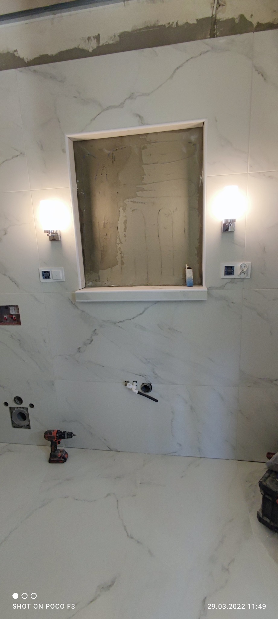 Niedokończona wnęka w ścianie łazienki z marmurowymi płytkami, widoczne instalacje hydrauliczne, wiertarka akumulatorowa i lampy ścienne.