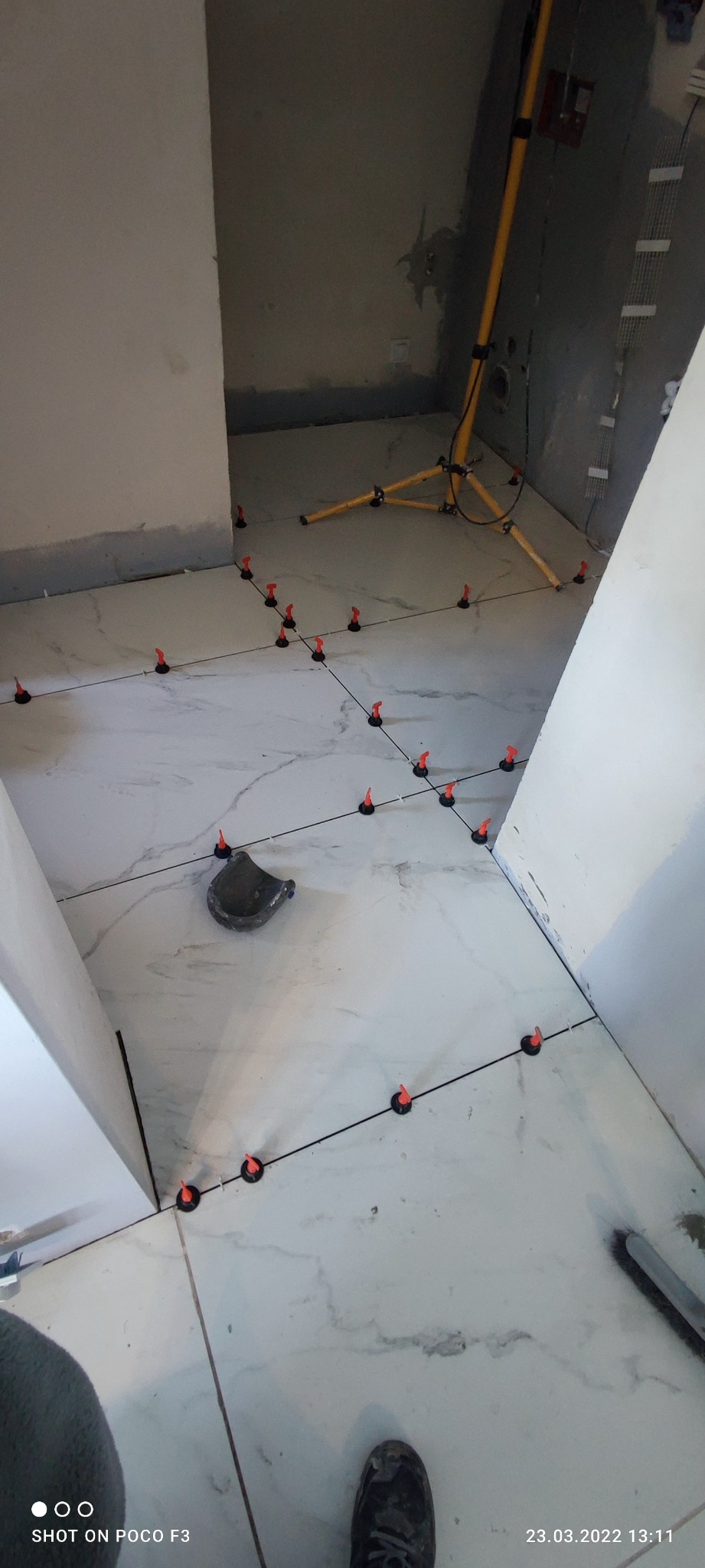 Układanie marmuropodobnych płytek podłogowych z użyciem systemu poziomowania, widoczna żółta lampa budowlana i szara ściana w tle.