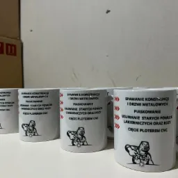 Rząd białych kubków z czarnym nadrukiem usług spawalniczych, piaskowania, usuwania starych powłok lakierniczych oraz cięcia ploterem CNC, z ilustracją spawacza.