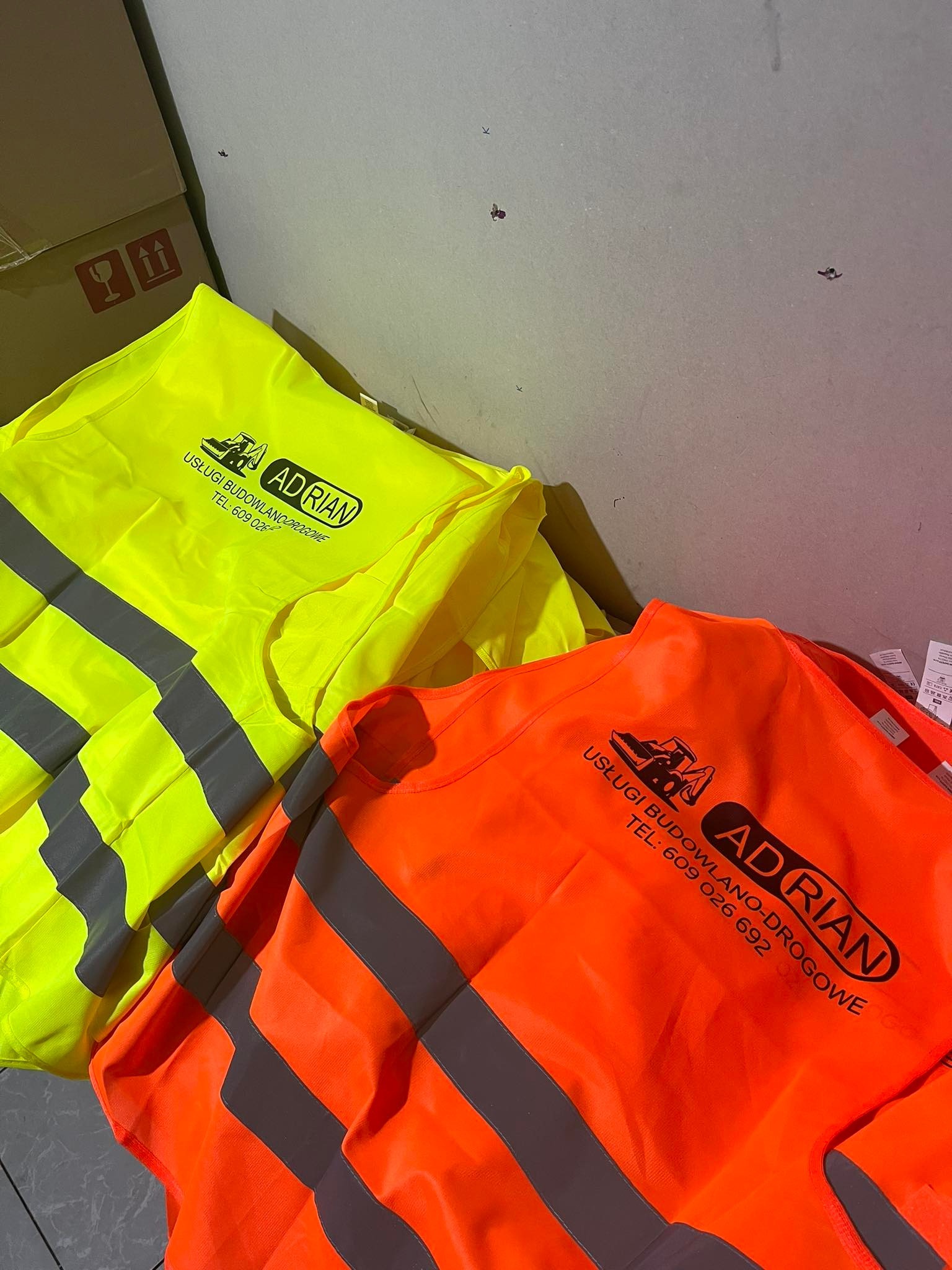 Dwa złożone bezrękawniki odblaskowe: żółty i pomarańczowy, z nadrukowanym logo firmy budowlanej i numerem telefonu.