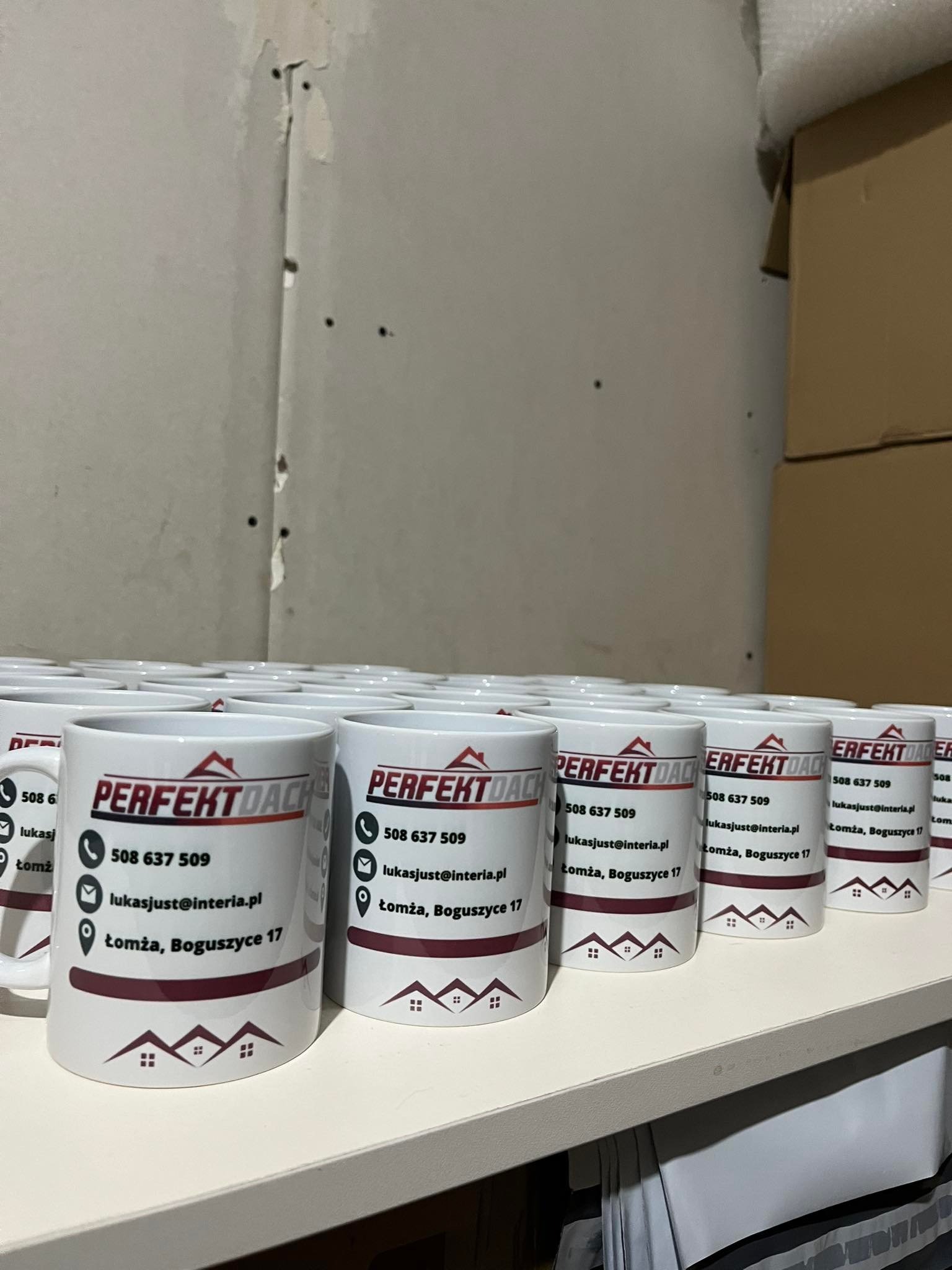 Rząd białych kubków z nadrukowanym logo firmy dekarskiej 'Perfekt Dach' wraz z danymi kontaktowymi, ustawionych na białej półce w magazynie.