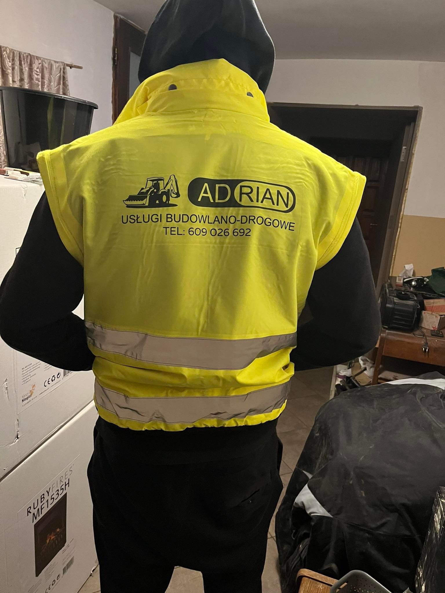 Żółta kamizelka odblaskowa z nadrukiem logo firmy budowlano-drogowej 'Adrian', widoczna z tyłu na osobie w czarnej bluzie z kapturem, w tle wnętrze pomieszczenia z pudełkami i sprzętem.