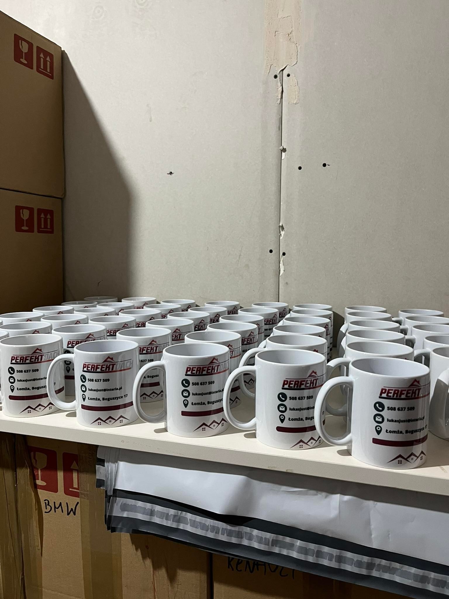 Rząd białych kubków z nadrukowanym logo firmy 'Perfekt Dach' i danymi kontaktowymi, ustawionych na białej półce na tle surowej ściany i kartonów z oznaczeniami ostrzegawczymi.