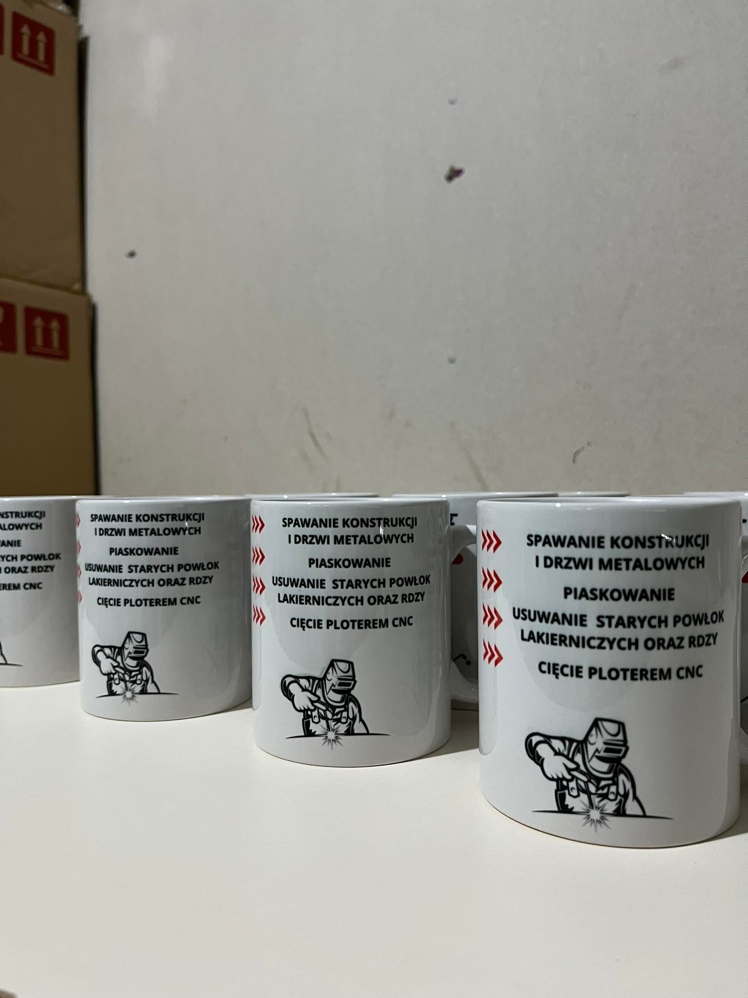 Rząd białych kubków z czarnym nadrukiem usług spawalniczych, piaskowania, usuwania starych powłok lakierniczych oraz cięcia ploterem CNC, z ilustracją spawacza.