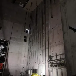 Wysokie, surowe ściany betonowe w trakcie budowy, z widocznymi stalowymi wspornikami i rusztowaniami, oświetlone punktowo, z logo firmy Eiffage na czerwonym banerze.