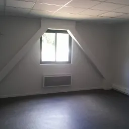 Puste poddasze z oknem w szczycie, szarą podłogą i białymi ścianami, widoczny grzejnik panelowy i listwy przypodłogowe.