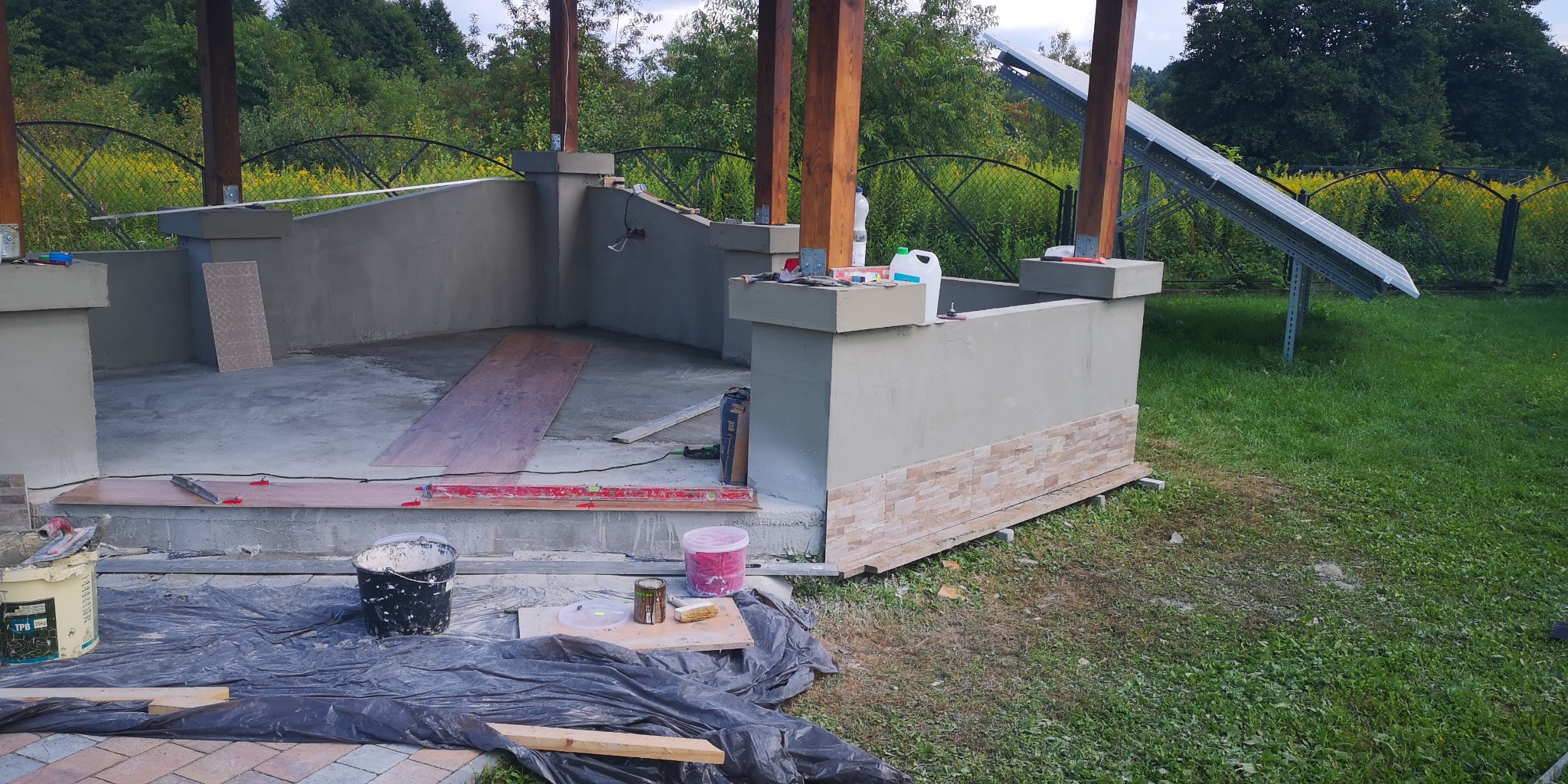 Budowa altany w ogrodzie: widać murowane ściany do połowy wysokości, drewniane słupy, częściowo ułożoną podłogę z desek, fragment paneli słonecznych w tle, wiaderka z zaprawą i narzędzia na folii...