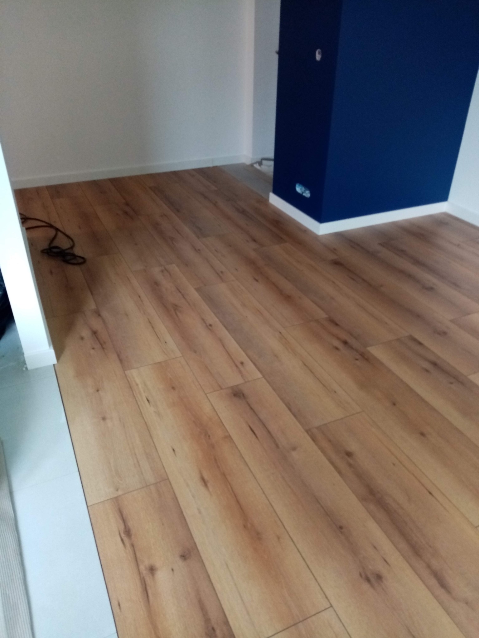 Ułożony parkiet laminowany w odcieniach brązu, z widoczną ciemnoniebieską kolumną i białymi listwami przypodłogowymi, kabel elektryczny leżący w rogu.