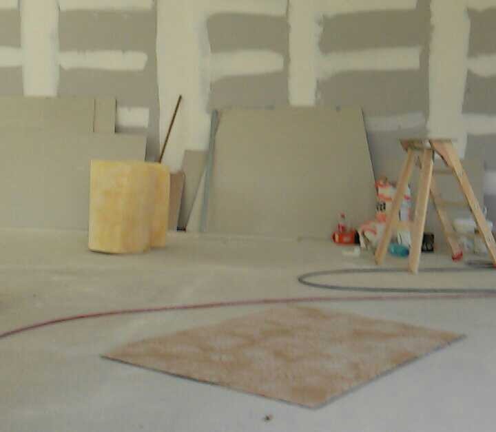 Wnętrze w trakcie prac wykończeniowych z widocznymi płytami kartonowo-gipsowymi na ścianach, rolką wełny mineralnej, drewnianą drabiną i zabezpieczonym fragmentem podłogi.