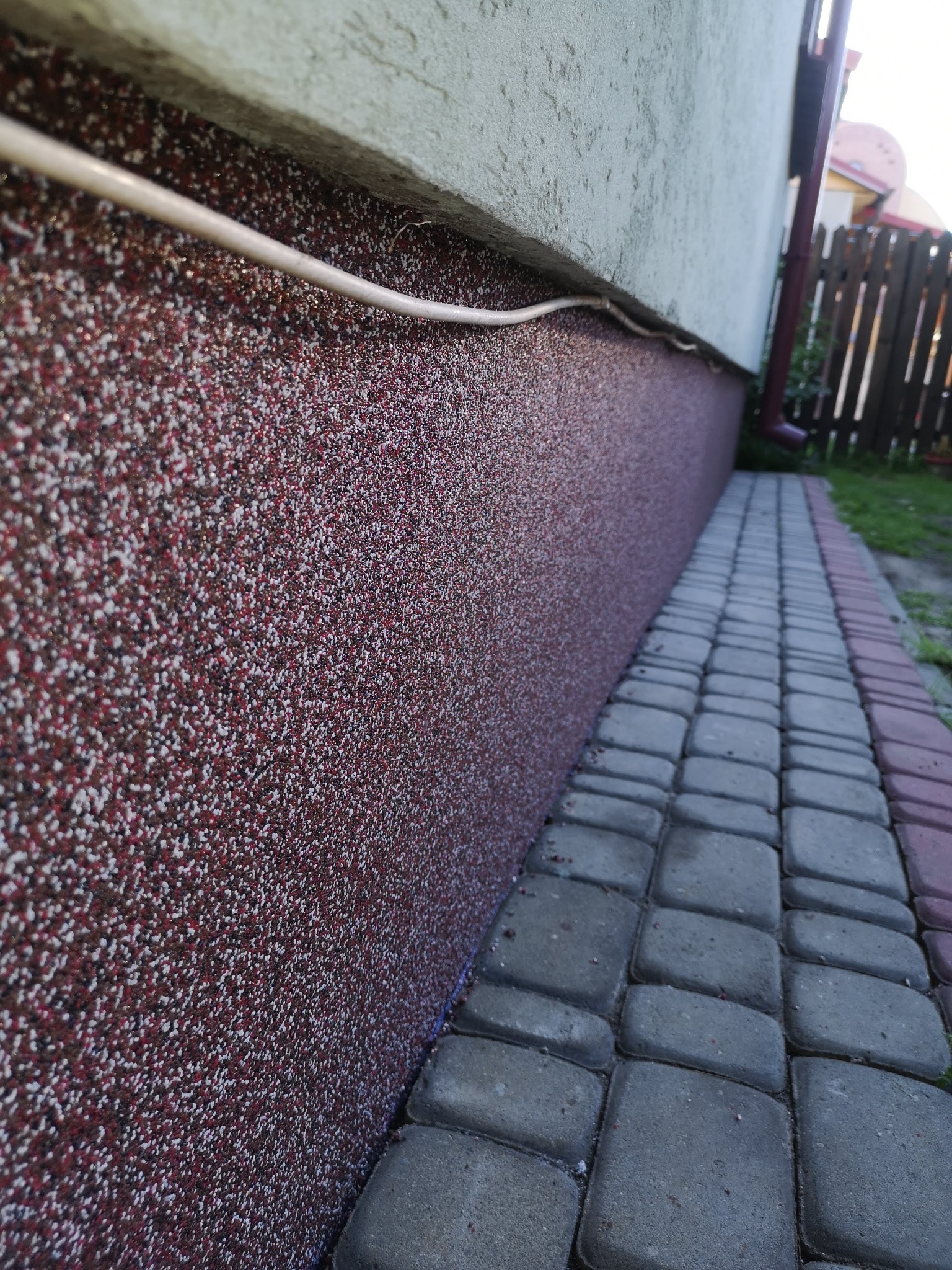 Fragment elewacji budynku z nałożonym tynkiem mozaikowym w odcieniach czerwieni, bieli i brązu, widoczny przewód elektryczny oraz fragment chodnika z kostki brukowej.