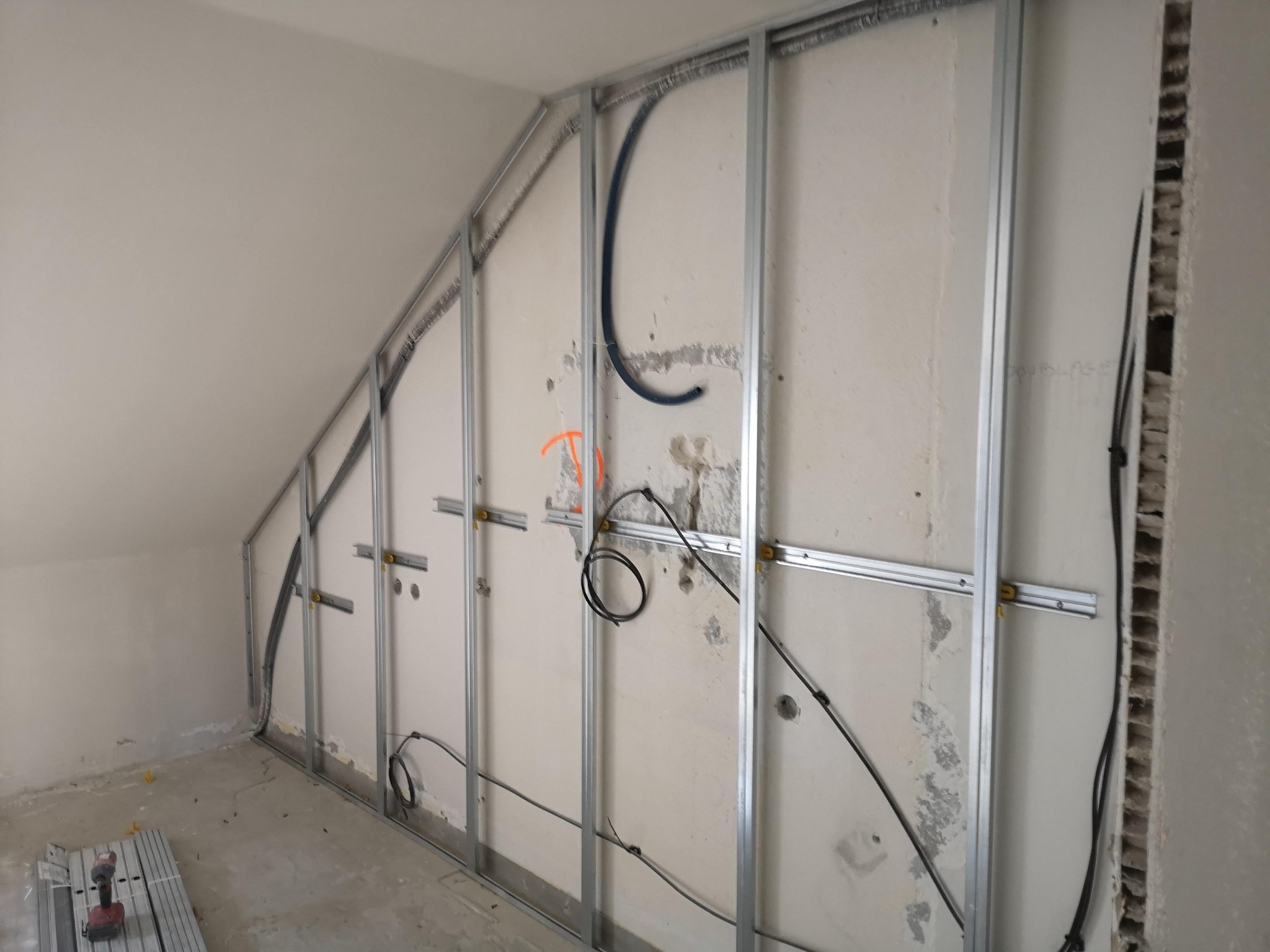 Skośny pokój na poddaszu w trakcie prac wykończeniowych: widoczne metalowe profile konstrukcyjne na ścianie, kable elektryczne i rury prowadzone wewnątrz, ślady po usuniętej starej powłoce ściennej...