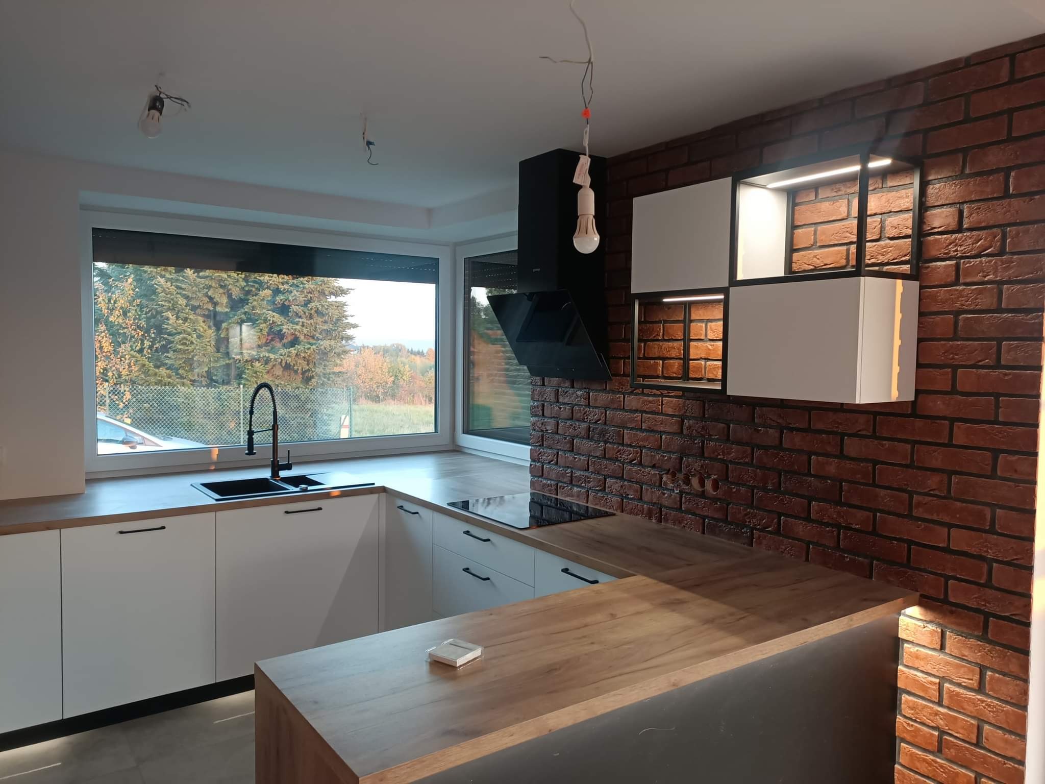Nowoczesna kuchnia z białymi szafkami, drewnianym blatem i ścianą z cegły, okno z widokiem na drzewa, czarny okap i niedokończone oświetlenie sufitowe.