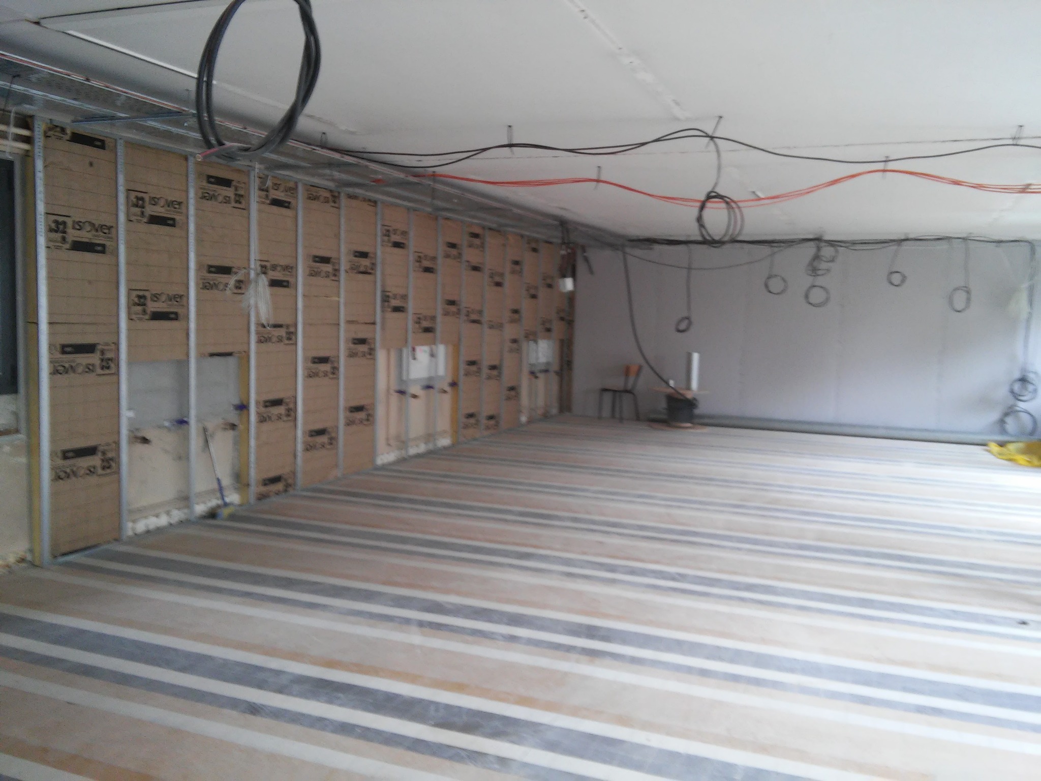 Wnętrze w trakcie prac wykończeniowych: widoczne metalowe profile konstrukcyjne, płyty izolacyjne z nadrukowanym logo producenta, częściowo położona podłoga z paskami w różnych odcieniach szarości...