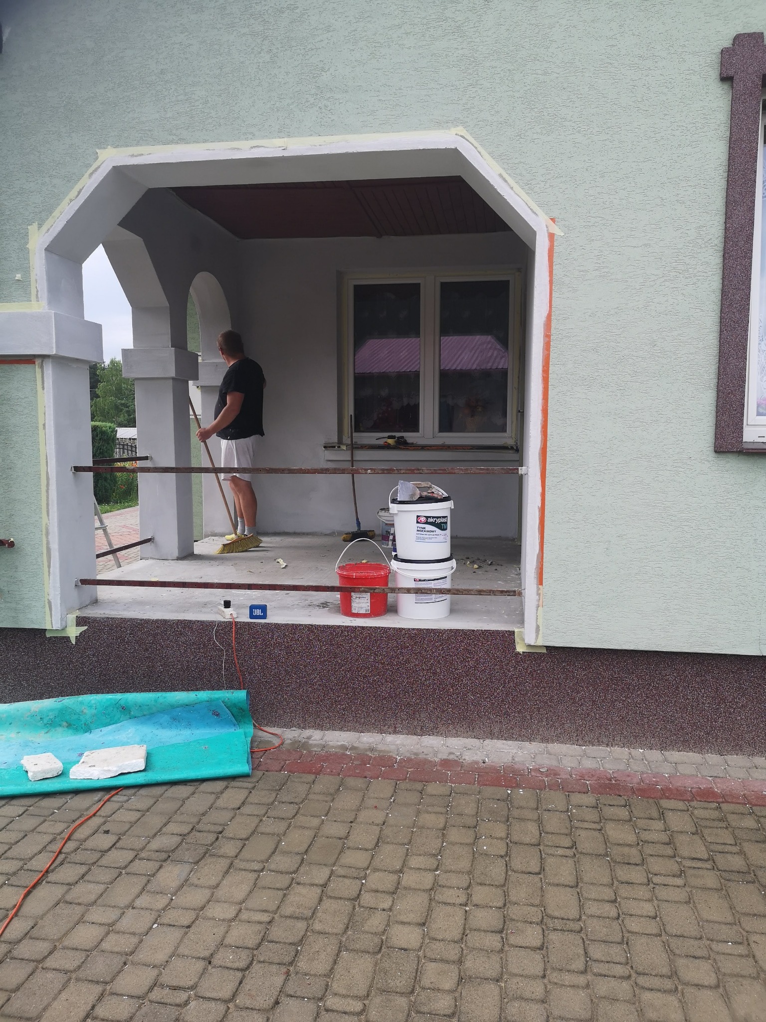 Malowanie elewacji domu z widocznym zadaszonym wejściem, przygotowane wiadra z farbą Akryplast, osoba zamiata podłogę, zabezpieczone taśmą malarską krawędzie.