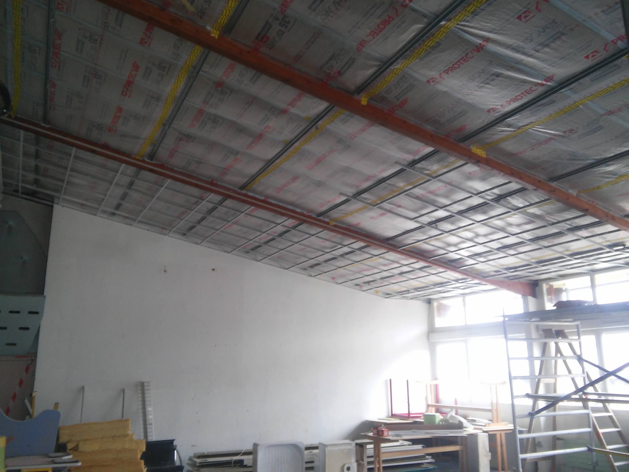 Montaż metalowej konstrukcji pod sufitem z izolacją termiczną, widoczna biała ściana i rusztowanie w pomieszczeniu z oknami.