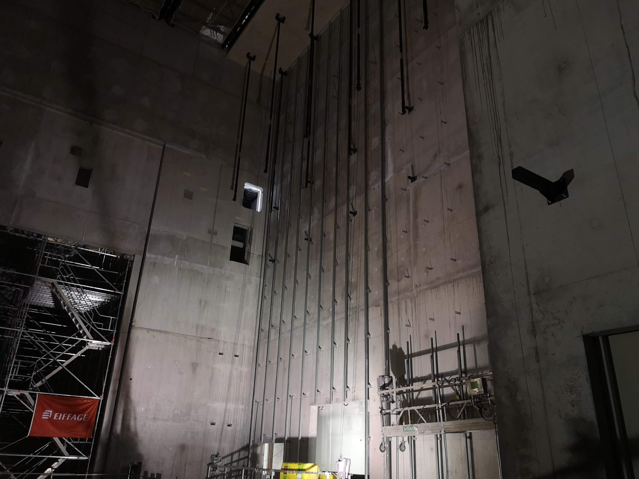 Wysokie, surowe ściany betonowe w trakcie budowy, z widocznymi stalowymi wspornikami i rusztowaniami, oświetlone punktowo, z logo firmy Eiffage na czerwonym banerze.