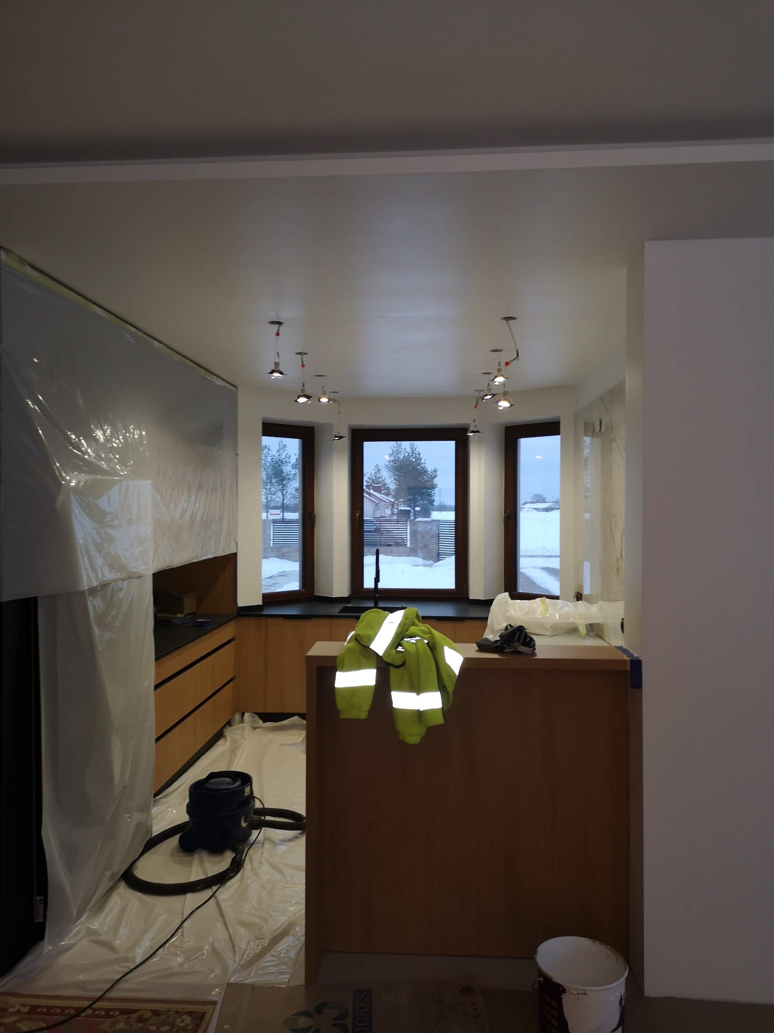 Wnętrze kuchni w trakcie prac wykończeniowych, zabezpieczone folią malarską, widoczny odkurzacz, ubranie robocze i wiadro z farbą.