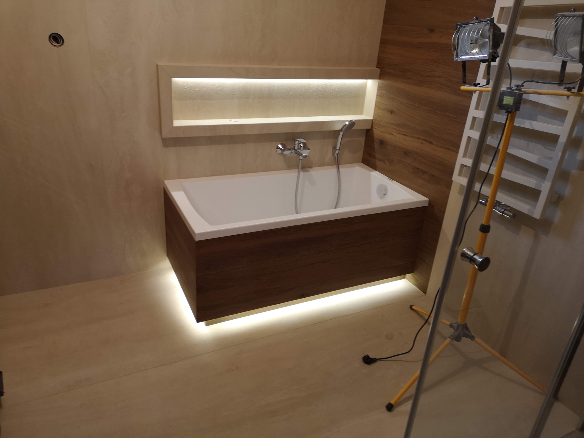 Nowoczesna wanna z obudową z ciemnego drewna, podświetlona od dołu, z zamontowaną baterią i słuchawką prysznicową, oświetlona lampami budowlanymi, w trakcie prac wykończeniowych.