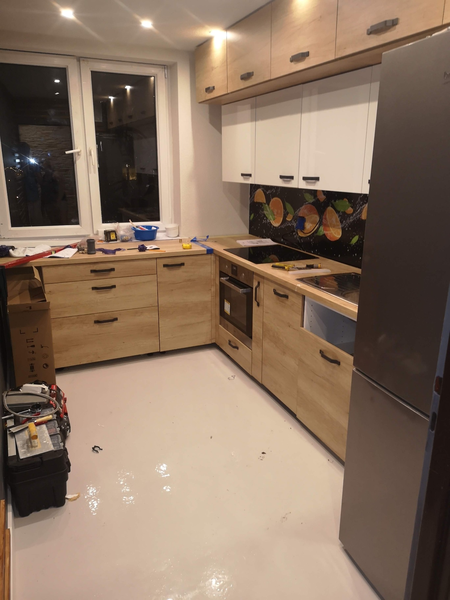 Montaż kuchni z jasnymi drewnianymi frontami, białymi szafkami wiszącymi i panelem z motywem pomarańczy, widoczne narzędzia i akcesoria montażowe, podłoga w trakcie wykańczania.