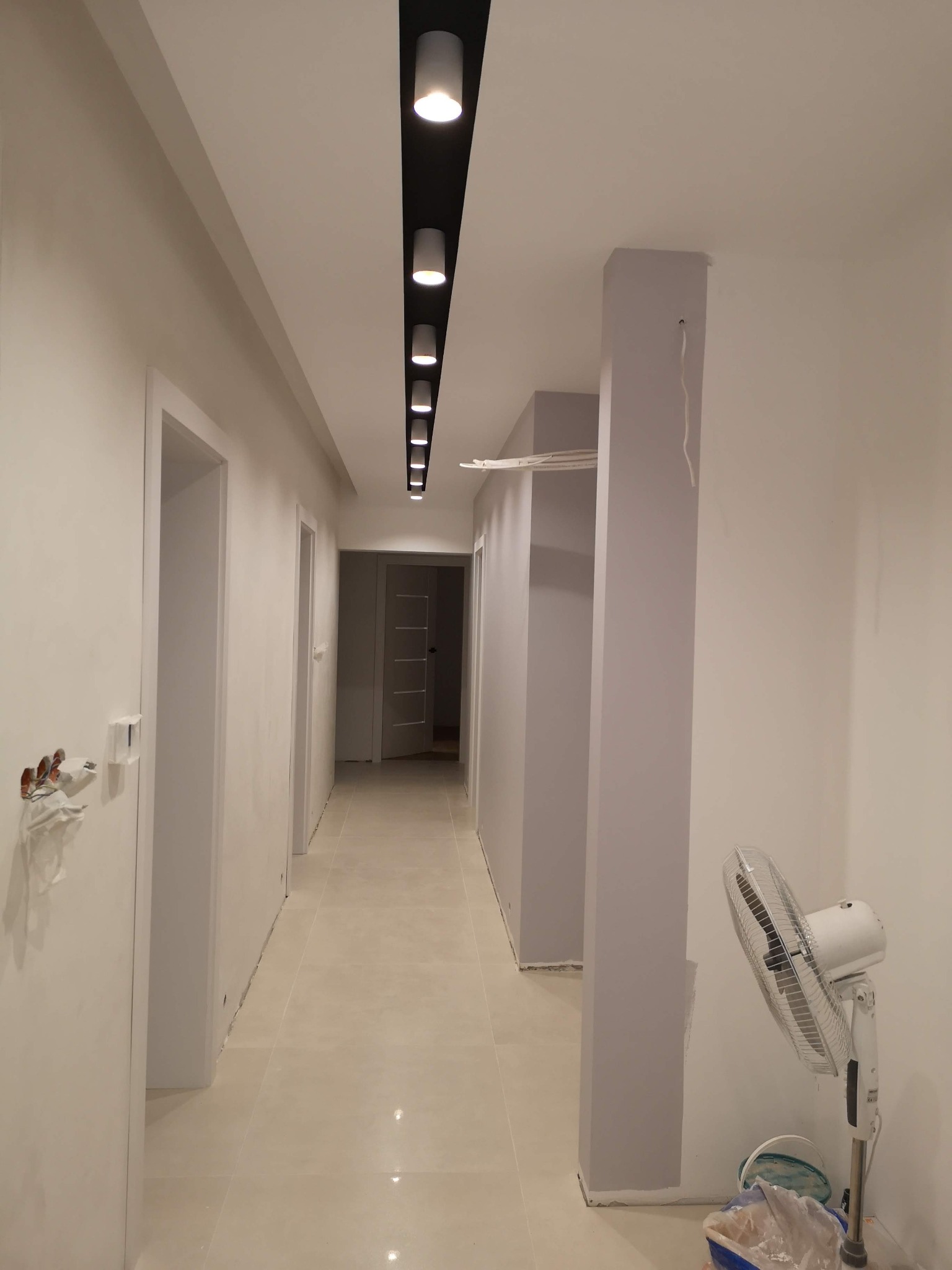 Korytarz w trakcie prac wykończeniowych: widoczne białe ściany, jasna podłoga z płytek, zamontowane drzwi, rząd reflektorów sufitowych oraz wentylator stojący i wiadro z farbą.
